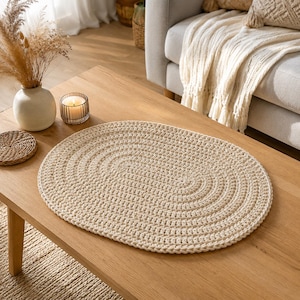 May include: An oval-shaped, cream-coloured crocheted placemat featuring a thick, textured yarn construction with concentric circular stitching patterns. The placemat is laid flat on a smooth, light-toned wooden surface, showcasing a uniform weave.