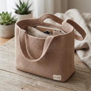 Könnte beinhalten: Beige gehäkelte Tragetasche mit zwei Henkeln, gefüllt mit einer beigen Reißverschlusstasche, einem Notizbuch und anderen Gegenständen. Die Tasche steht auf einer Holzoberfläche. Das Etikett "VivCrochets" ist sichtbar.