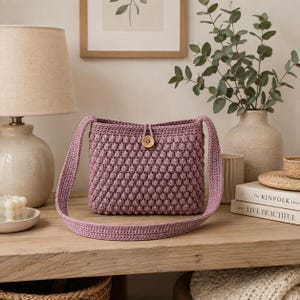 May include: A mauve crochet crossbody bag featuring a textured bobble stitch pattern, a single long shoulder strap, and a circular wooden button closure at the top center. The bag is constructed with a dense, uniform knit design.