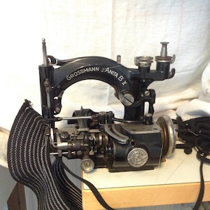 Puede incluir: Una máquina de coser negra vintage, "Grossmann Anita B II", con una tela a rayas blancas y negras adjunta. La máquina está sobre una mesa de color claro, con una cuerda negra cerca. La máquina tiene un emblema plateado.