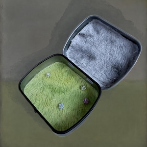 May include: A square metal container with an open lid, featuring a soft, lime green faux fur lining in the base and a grey faux fur lining in the lid. Four small, decorative flower-shaped pins are embedded into the green fur base.