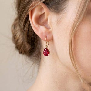 May include: A gold drop earring featuring a faceted, pear-shaped red gemstone suspended from a delicate gold hook. The earring hangs from an earlobe, showcasing the vibrant red color of the stone and the polished gold metal setting.