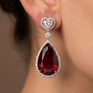 Op de afbeelding: Elegante oorhangers met een hartvormige oorbel met een halo van kleine diamanten. Een sprankelende ronde diamant verbindt met een grote, peervormige, dieprode edelsteen, ook omgeven door een diamanten halo. Sieraden.