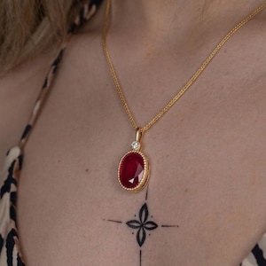May include: A gold chain necklace featuring an oval-shaped red gemstone pendant. The pendant is framed by a beaded gold border and topped with a small, round, clear white stone. The necklace rests against the skin, showcasing the metal link chain.