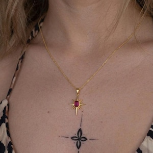 May include: A gold-toned necklace featuring a delicate chain and a pendant shaped like a starburst with a rectangular, deep pink gemstone at its center. The pendant is suspended from a thin, polished gold-colored metal chain that rests on the neck.