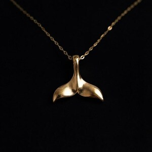 Op de afbeelding: Een goudkleurige ketting met een hanger in de vorm van een walvisstaart. De hanger heeft een glad, reflecterend oppervlak en hangt aan een fijne, dunne goudkleurige ketting. Het ontwerp is eenvoudig en richt zich op de gebogen vorm.