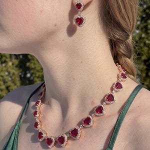 Op de afbeelding: Een sieradenset bestaande uit een ketting en hangende oorbellen. Elk sieraad bevat hartvormige rode stenen omringd door kleine heldere steentjes, gezet in een roségoudkleurig metalen frame. De ketting bestaat uit meerdere schakels.
