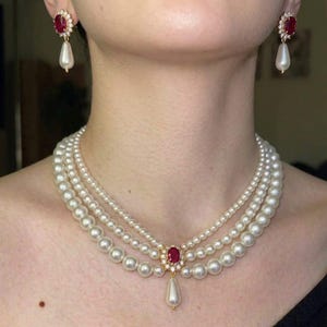 May include: A three-strand pearl necklace featuring a central oval red gemstone surrounded by small clear stones and a single dangling teardrop pearl. Matching earrings consist of an oval red gemstone with a dangling teardrop pearl.