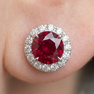 May include: A round stud earring featuring a large, deep red faceted ruby at the center, secured by four silver-toned prongs. The ruby is surrounded by a halo of small, clear, round-cut white diamonds set in a circular silver-toned metal frame.