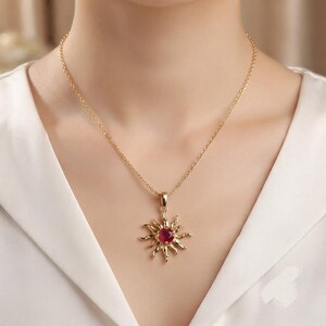 May include: A gold-toned necklace featuring a sun-shaped pendant with a round, deep red gemstone at its centre. The pendant is suspended from a delicate gold-toned chain, resting against a plain white fabric background.
