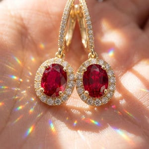 May include: A pair of gold drop earrings featuring oval-cut red gemstones surrounded by a halo of small white diamonds. The earrings have a hinged hoop mechanism, with additional small diamonds set along the front of the gold hoop structure.