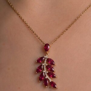 May include: A gold necklace featuring a pendant shaped like a leaf or branch. The pendant is adorned with multiple marquise-cut deep red rubies and small clear accent stones set along the central stem, suspended from a delicate gold chain.