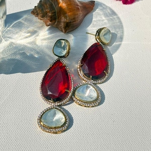 May include: A pair of drop earrings featuring large, faceted, pear-shaped deep red stones and smaller, clear, rounded stones. Each stone is set in a gold-toned metal frame bordered by tiny, sparkling white crystals, creating a layered, dangling design.