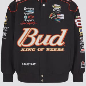 Puede incluir: Chaqueta bomber negra con los logotipos de Budweiser y Goodyear, y varios parches de patrocinadores. La chaqueta presenta detalles en rojo y blanco, cierre de botones y cuello alzado. El texto "Bud King of Beers" se muestra de forma destacada.