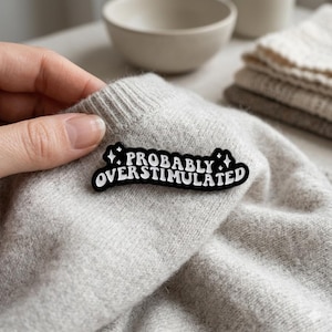 Peut inclure: Une épingle en émail noir et blanc avec le texte "PROBABLY OVERSTIMULATED" est affichée sur un pull gris clair. L'épingle a une bordure noire et des lettres blanches. Le pull est plié, avec d'autres articles en arrière-plan.
