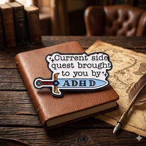 May include: A pin with the text "Current side quest brought to you by ADHD" in white, black, and blue. The pin is shaped like a sword and is placed on a brown leather-bound book. A map and quill are also visible.
