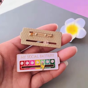 Può includere: Una spilla rettangolare in smalto con la scritta "MY SOCIAL BATTERY" e un indicatore del livello della batteria con faccine emoji. La spilla ha un retro color oro con una chiusura a spilla da balia.