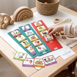 Könnte beinhalten: Ein Kind interagiert mit Aktivitätskarten auf einem weißen Tisch. Die Karten befinden sich auf roten und türkisfarbenen Kartenhaltern. Die Karten zeigen Illustrationen von täglichen Aktivitäten wie Wäsche waschen, spielen und zur Schule gehen. Holzspielzeug und ein Weidenkorb befinden sich ebenfalls auf dem Tisch.
