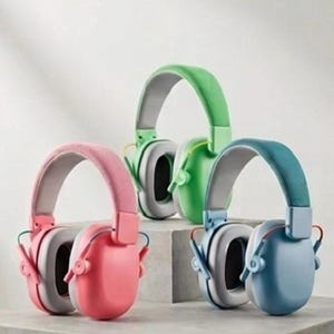 May include: Three pairs of over-ear headphones in pink, green, and blue. Each pair has a padded headband and earcups in a matching colour, with grey accents. The headphones are displayed on a grey surface. The design is modern and minimalist.