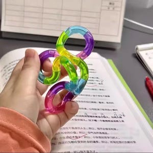 May include: A colorful tangle toy in shades of pink, purple, blue, green, and yellow. The plastic toy is held in a hand and appears to be made of interlocking curved segments. The background includes a book and a pen.