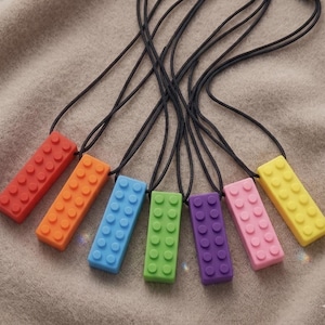 Puede incluir: Una colección de siete colgantes coloridos en forma de ladrillo sobre cordones negros. Los colgantes son rojos, naranjas, azules, verdes, morados, rosas y amarillos. Están colocados sobre una superficie texturizada de color neutro.