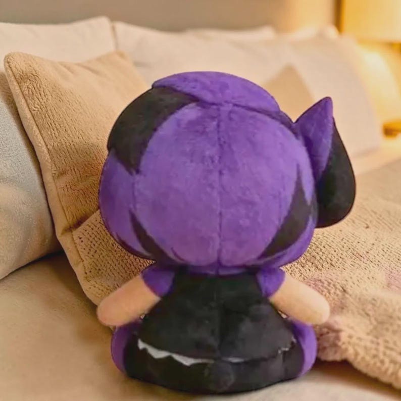 May include: A plush doll with a purple head and black details, wearing a black dress. The doll has tan arms and is sitting on a bed with pillows.
