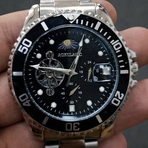 Puede incluir: Un reloj de pulsera de acero inoxidable en tono plateado con esfera negra y bisel giratorio. La esfera muestra el texto AQKULASIC, un indicador de fase lunar, un volante esqueleto y una ventana de fecha con una lente de aumento.