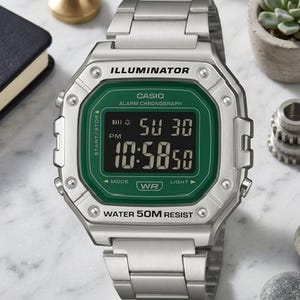Puede incluir: Un reloj digital Casio plateado con pantalla verde. La esfera del reloj muestra la hora y la fecha. La correa del reloj es de metal plateado. El reloj tiene la palabra "ILLUMINATOR" en la parte superior y "WATER 50M RESIST" en la inferior.
