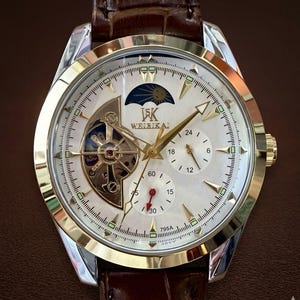 May include: A gold and silver wristwatch with a white face, featuring a complex dial design with multiple sub-dials and a visible mechanical movement. The watch has gold hands and markers, a brown leather strap, and the text "WELEIKAI".