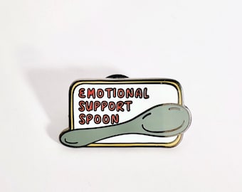 May include: A white enamel pin with a black outline. The pin features a cartoon spoon with the text "Emotional Support Spoon" in red and black.