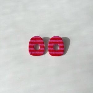 Può includere: Due orecchini ovali con un motivo a righe nei toni del rosso e del rosa. Ogni orecchino presenta un foro centrale ed è posto su uno sfondo bianco. Questi orecchini sono un accessorio di moda.