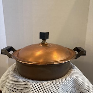 May include: A copper cooking pot with a lid and two handles. The lid has a black square knob. The pot is sitting on a white lace tablecloth. The pot is made of copper and has a shiny finish.