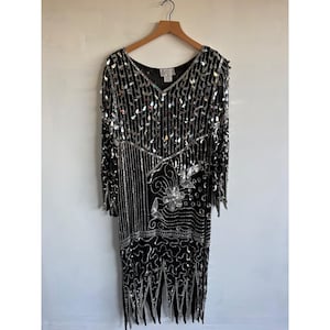 May include: A black long-sleeve dress featuring intricate silver sequin and bead embellishments in teardrop, floral, and swirling patterns. The garment has a V-neckline, long sleeves, and a hemline with pointed, dangling beaded fringe details.