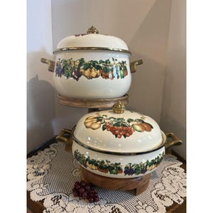 Puede incluir: Dos ollas de esmalte color crema con tapas, que presentan un patrón de frutas coloridas en los lados. Cada olla tiene dos asas de metal en tono dorado y un pomo a juego en la tapa. El diseño de frutas incluye uvas, peras y cerezas.