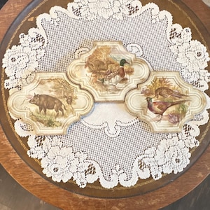 May include: Three decorative ceramic tiles with wildlife scenes. One tile depicts a wild boar, another shows ducks, and the third features pheasants. The tiles have a cream-colored background and are arranged on a round table with a lace doily.