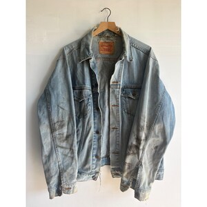Könnte beinhalten: Eine hellblaue Jeansjacke mit Used-Look, ausgestattet mit einem spitzen Kragen, langen Ärmeln mit geknöpften Manschetten, zwei Brusttaschen mit geknöpften Klappen und einer zentralen Knopfleiste. Ein braunes Lederetikett von Levi Strauss & Co.