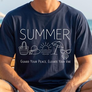 May include: Navy blue t-shirt with the word "SUMMER" and summer-themed illustrations in white. The shirt also features the text "Guard Your Peace, Elevate Your Vibe". The person is sitting on a wooden beach chair.