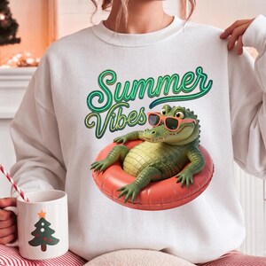 May include: White sweatshirt with a graphic of a green alligator wearing sunglasses and floating in a red inner tube. The words "Summer Vibes" are written above the alligator in green and yellow script.