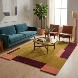 May include: A rectangular area rug featuring a large mustard yellow center with geometric blocks of deep burgundy and soft peach at the edges. The rug is placed in a living room setting with a teal sofa, two brown chairs, and a round glass coffee table.