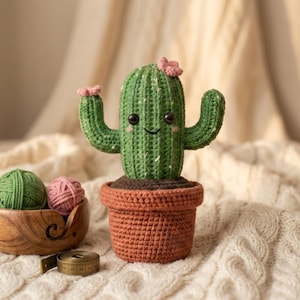 May include: A handcrafted crochet cactus with a smiling face and pink flower accents, potted in a terracotta-colored pot. The green cactus has two arms and is set against a soft, neutral background. Yarn balls and a measuring tape are also visible.
