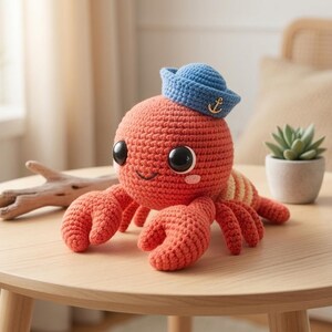 Può includere: Un giocattolo a forma di granchio lavorato all'uncinetto in arancione con grandi occhi neri e guance rosa, che indossa un piccolo cappello da marinaio blu decorato con un emblema di ancora dorata. Il granchio ha due grandi chele anteriori e diverse zampe più piccole, con un motivo a righe sul dorso.