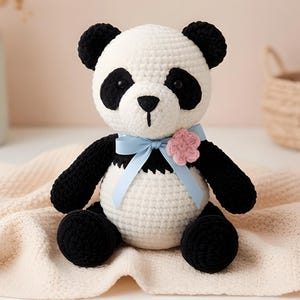 May include: A handmade crochet panda bear with black and white coloring. The panda has black eyes, ears, and limbs, with a white body and head. A light blue ribbon and pink flower adorn the panda's neck. The panda is sitting on a cream-colored blanket.