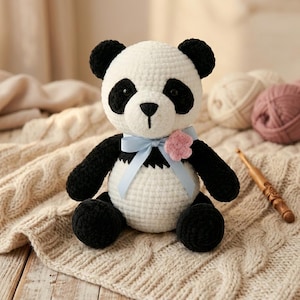 Könnte beinhalten: Handgehäkelter Panda-Bär aus schwarzem und weißem Garn. Der Panda hat schwarze Augen, Ohren und Gliedmaßen und einen weißen Körper. Ein hellblaues Band und eine rosa Blume schmücken seinen Hals. Der Panda sitzt auf einer cremefarbenen Strickdecke.