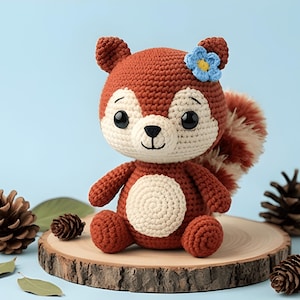 May include: A handmade crochet squirrel doll with a brown body, cream-colored face and belly, and a fluffy tail. The squirrel has a blue flower on its head and is sitting on a wooden slice. The background is a light blue.