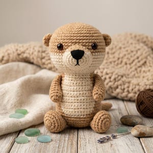 May include: A small, handcrafted crochet otter toy featuring a light tan and cream-coloured yarn body. The otter has two dark brown safety eyes, a black embroidered nose, and a rounded head with small ears, sitting in an upright position.
