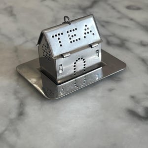 May include: A silver-coloured metal tea infuser shaped like a small house with a perforated roof spelling the word TEA. The house features a small metal loop at the top and sits on a matching rectangular metal drip tray with a reflective surface.