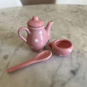 May include: A miniature pink ceramic tea set consisting of a teapot with a removable lid, a small teacup, and a spoon. Each piece features a glossy finish and is decorated with white polka dots, creating a cohesive and charming set of dollhouse accessories.