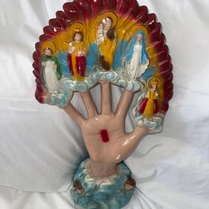 May include: A religious figurine depicting a hand with figures of saints and religious figures. The hand is painted in a flesh tone, with a red mark on the palm. The figures are in various colors, set against a red, yellow, and blue backdrop.