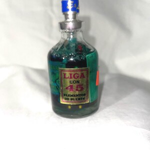 May include: A small clear glass bottle filled with a dark green liquid. The bottle features a rectangular label with a gold border and red text that reads LIGA CON 45 ELEMENTOS DE SUERTE. It has a silver neck and a blue cap with gold lettering.
