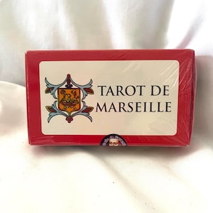 May include: A red tarot card deck box with the words "TAROT DE MARSEILLE" in black text. The box has a white label with a colorful emblem. The deck is wrapped in clear plastic.